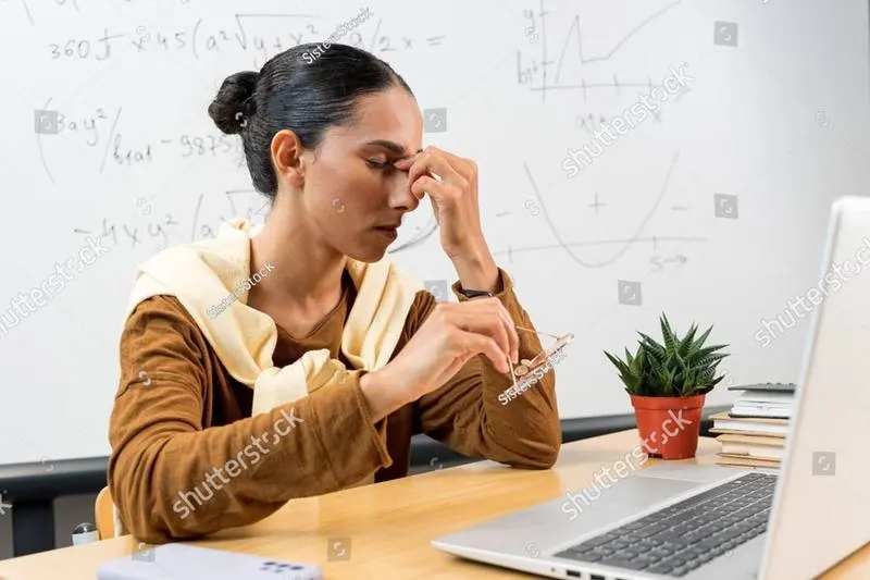 A woman at a desk with her glasses off, pinching the bridge of her nose