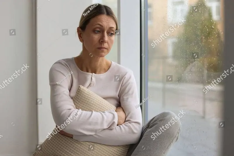A woman sitting in a window seat holding a cushion, looking away, rain on the glass